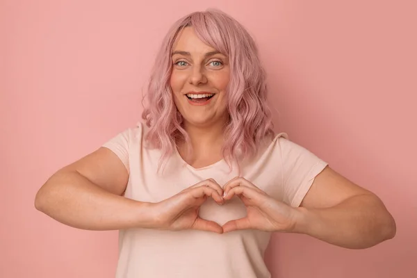 可愛くて面白いピンクの髪の女性若いです女性は幸せとロマンチックな作るハート形のポーズに対してピンクの背景 — ストック写真