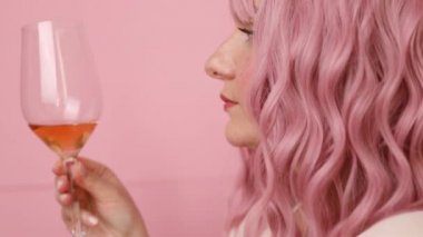 Güzel, rahat, pembe kıvırcık saçlı, gül şarabı içen, şarap kadehini parlak pembe arka planda tutan