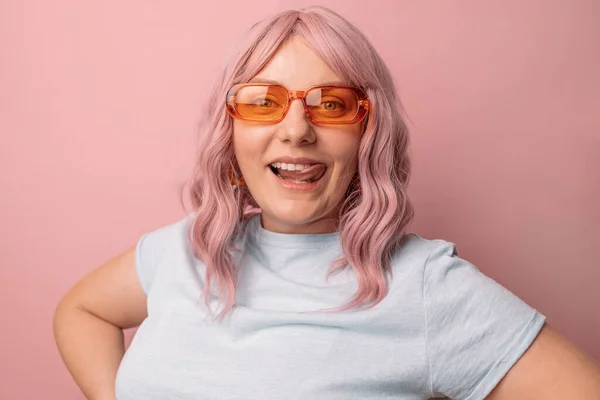 幸せな若いです30代の女性ブロガーで明るいオレンジスタイリッシュな眼鏡でカメラを見て 彼の舌と美しい白い歯を示していますピンクの背景に孤立 — ストック写真