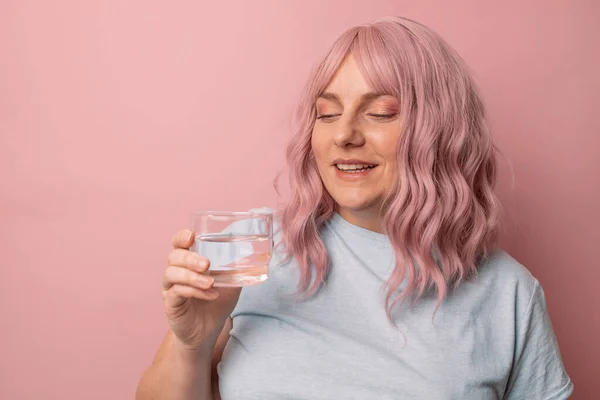 ピンクの背景に隔離された水のガラスを保持しながら 美しい若い女性の笑顔 — ストック写真