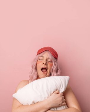 Güzel, genç ve güzel bir kadın, elinde yumuşak bir yastıkla uyku maskesi takıyor, uyumak istercesine esniyor, pembe arka planda yalnız bir şekilde sabahın köründe uyandığı için üzgün..