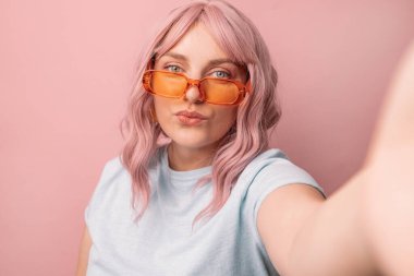 Parlak turuncu gözlüklü göz alıcı 30 'lu yılların kadın blogcusu kameraya bakar ve pembe arka planda selfie çeker. Halk konsepti