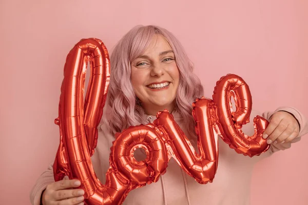 ピンクの背景に赤い箔の愛の言葉風船を持つ楽しさを持つかなり若い20代の女性の肖像画。バレンタインデー母の日誕生日. — ストック写真