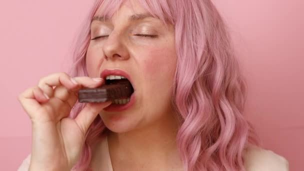 Νεαρή γυναίκα δαγκώνει ένα κομμάτι σοκολατένιας καραμέλας που απομονώνεται σε ροζ φόντο. — Αρχείο Βίντεο