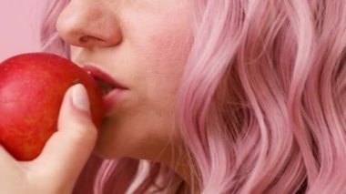 Çekici pembe saçlı kadın pembe arka planda elma yiyor.