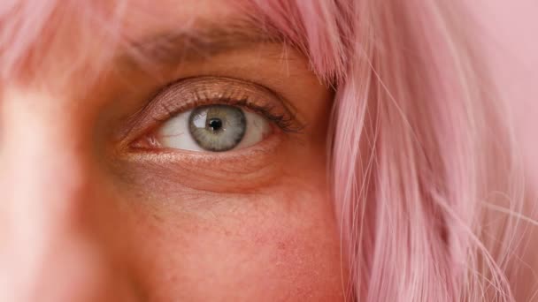 Primer plano del ojo humano femenino con pelo rosa. Mujer con maquillaje de belleza facial natural aislado sobre fondo rosa — Vídeos de Stock