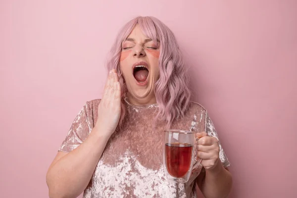眠い疲れたピンクの髪の女性は口をカバーし、 yawnsはピンクの背景に対して立つ。朝お茶を飲む若い女の子. — ストック写真