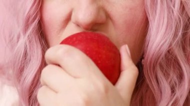 Pembe arka planda elma yiyen genç, pembe saçlı kadının yakın çekimi.