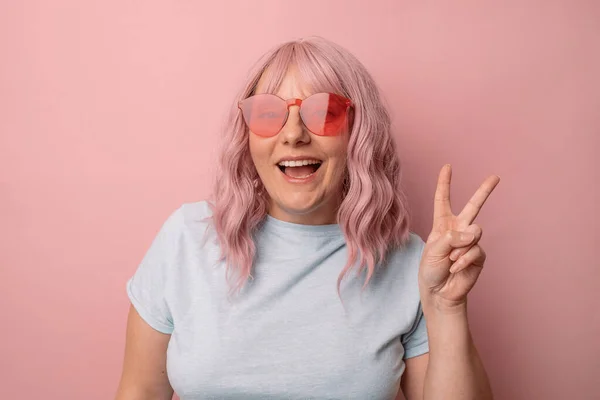Szczęśliwa kobieta w jasnych stylowych okularach przeciwsłonecznych sprawia, że znak V pokazuje gest pokoju uśmiechnięty i pozowanie izolowane na różowym tle studio. — Zdjęcie stockowe