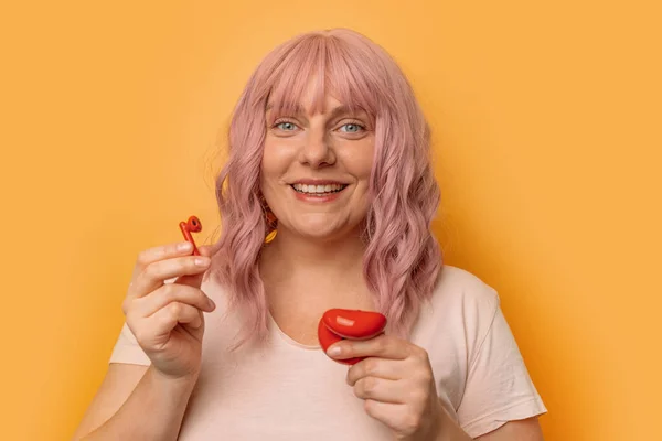 분홍색 곱슬머리에 노란색 배경에 고립된 귀에 무선 이어폰을 삽입하는 매력적 인 코카서스 여성의 모습. — 스톡 사진