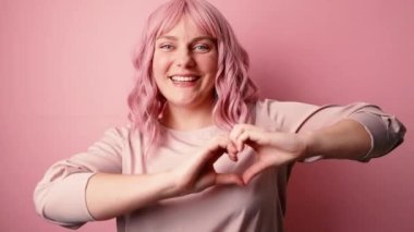 Güzel bir genç kız pembe arka planda izole edilmiş kameraya bakarak sevgiyi simgeleyen parmaklarıyla kalbini gösteriyor.