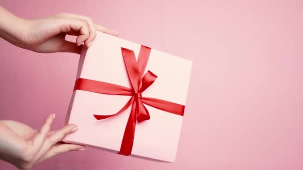 Mão branca mostrando caixa de presente surpresa com laço de fita vermelha isolada na parede de pastel rosa — Vídeo de Stock