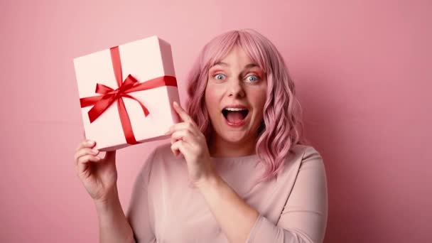 Celebración, feliz positiva emocionada mujer caucásica divertida sosteniendo presente caja de regalo con lazo de cinta roja posando aislado sobre fondo rosa. Estudio — Vídeos de Stock