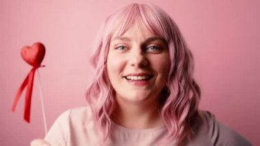 Romantik, mutlu, romantik kalp dekoru olan beyaz bir kadın pembe arka plandaki kameraya bakıyor. Sevgi. Sevgililer Günü. Duygular.