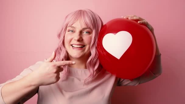 Positive nice young happy woman in good mood smiling holding red balloon shaped heart isolated on pink background — Stock Video