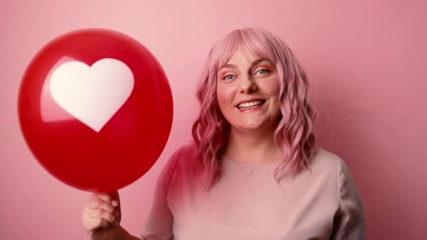 Ung söt kaukasisk kvinna ler håller röd ballong format hjärta isolerad på rosa bakgrund. Alla hjärtans dag firande — Stockvideo