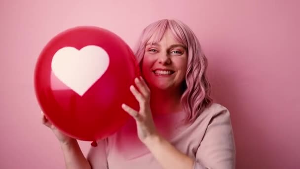 Pozitivní pěkný mladý šťastný s červeným srdcem vzduchem balón dívá na kameru a usmívá se izolované růžové pozadí. Sdílej lásku. Valentýn. — Stock video
