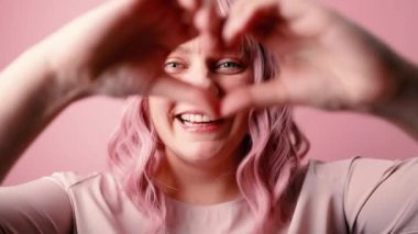 Güzel, 30 'lu yaşlarda beyaz, neşeli bir kadın gülümsüyor ve pembe arka planda elleri izole bir kalp şekli gösteriyor.