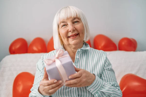 Feliz 50 años de edad, señora rubia dando caja de regalo en la cámara el 14 de febrero sentado en un sofá en casa. —  Fotos de Stock