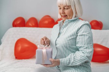 50 yaşında şık bir bluz giymiş sarışın bir kadın 14 Şubat 'ta evinde bir koltukta sürpriz bir hediye paketi açıyor..