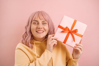 Mutlu, genç, heyecanlı, aşık beyaz kadın pembe arka planda izole edilmiş kırmızı kurdeleli hediye kutusunu tutuyor. Stüdyo