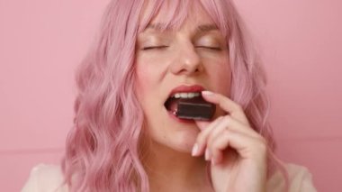 Pembe arka planda izole edilmiş çikolatalı waffle şekerini ısıran güzel bir genç kadının portresi.