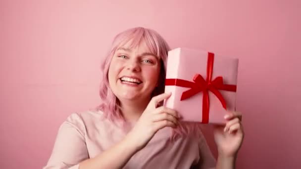 Счастливые молодые розовые волосы 30-х кавказская женщина в хорошем настроении проведения настоящее подарочная коробка с красной лентой бант позировать изолированы на розовом фоне — стоковое видео