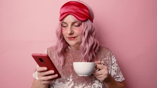 Portret van vrolijke slaperige vrouw draagt rood oogmasker en roze pyjama, maakt gebruik van mobiele telefoon aangesloten op draadloos internet drinkt koffie op gele achtergrond — Stockvideo