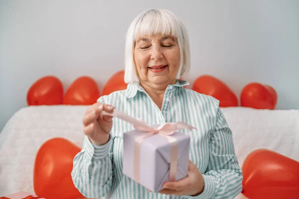 Mujer rubia de 50 años mayor en una elegante blusa desempaca una caja de regalo sorpresa el 14 de febrero sentada en un sofá en casa. Feliz día de San Valentín. —  Fotos de Stock