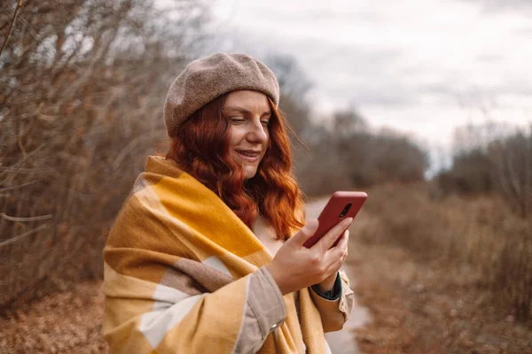 Güzel kızıl saçlı, gülümseyen bir kız internette sohbet etmek için akıllı telefonu kullanıyor ve şehir parkında rahatça yürüyor.