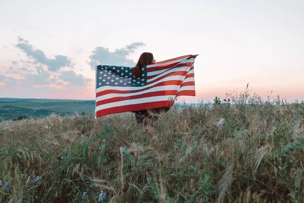Młoda kobieta dumnie trzyma amerykańską flagę na letnim polu. Patriota wznosi narodową amerykańską flagę przeciwko błękitnemu niebu. Dzień Niepodległości, 4 lipca. — Zdjęcie stockowe