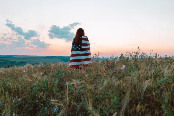 Młoda kobieta dumnie macha amerykańską flagą na letnim polu. Patriota podnosi amerykańską flagę przeciwko błękitnemu niebu. Dzień Niepodległości, 4 lipca. — Zdjęcie stockowe