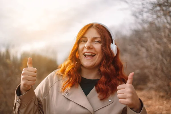 在城市公园里，身穿超大米色风衣的卷发女人一边听着智能手机和无线耳机的音乐，一边开心地笑着 — 图库照片