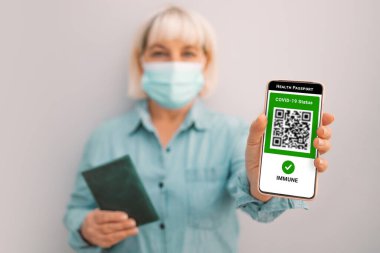 Covid-19 kontrol. 50 'li yaşlarda maskeli beyaz bir kadın elinde dijital yeşil kartı ve ekranında QR kodu olan akıllı bir telefon tutuyor. Sağlık pasaportu veya dokunulmazlık belgesi.