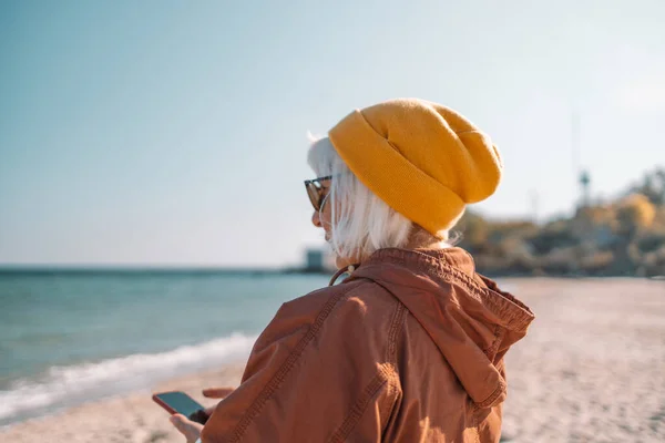 Atractiva aplicación de teléfono inteligente de mediana edad para mujer táctil que se relaja al aire libre en la playa de arena contra el mar —  Fotos de Stock