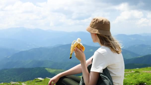 Az utazó felfedező lány banángyümölcsöt eszik a hegy tetején egy napos nyári napon. Siker, vándorlás és sport koncepció. — Stock videók