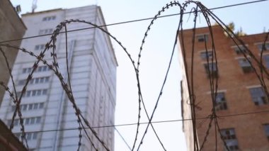 Dikenli tellerle çevrili yüksek beton çitler mavi gökyüzü arka planına karşı. Kırık camlı terk edilmiş tuğla binalar.