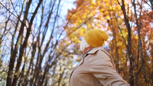 Šťastná padesátnice ve stylovém světlém oblečení s dlouhou žlutou šálou, která si na podzim vychutnává sama v lese s barevnými listy. Cestovní koncept. — Stock video