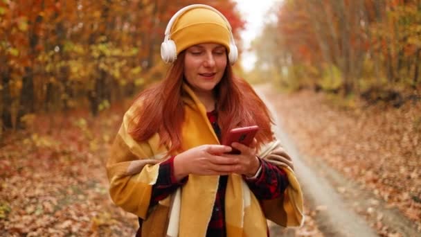 Actieve blanke vrouw wandelen samen met koptelefoon met behulp van mobiele telefoon geniet van de natuur in een stad herfst park. 5G-technologie apparaat. — Stockvideo