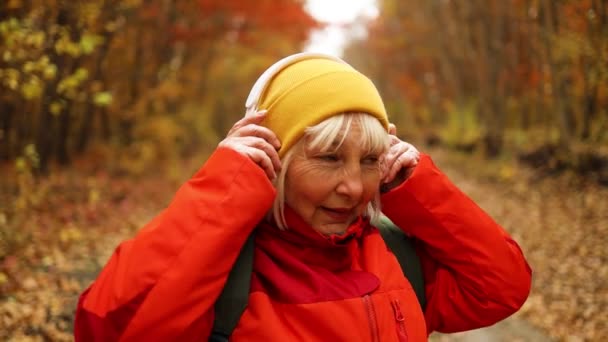 Glückliche Frau in bunten gelben Klamotten, die kabellose Kopfhörer aufsetzt, hört Musik beim Spaziergang im Herbst-Waldpark in der Natur — Stockvideo