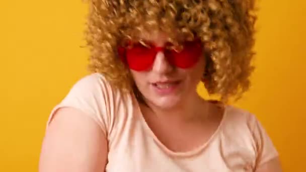 Glückliche junge Frau mit lockigem, flauschigem Haar in stylischer Liebes-Brille hebt die Arme und tanzt zu Lieblingsmusik isoliert vor gelbem Hintergrund — Stockvideo