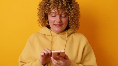 30 'ların çekici kadın blogcusu akıllı telefondan kameraya bakar ve sarı arka planda selfie çeker. Halk konsepti