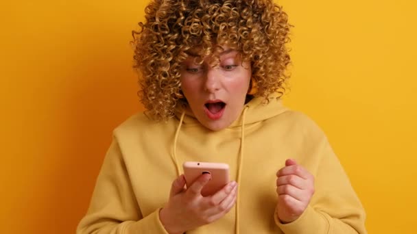 Retrato de una joven hermosa mujer caucásica sintiéndose feliz o sorprendida y sosteniendo el teléfono inteligente aislado sobre fondo amarillo — Vídeos de Stock