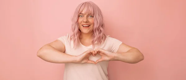 Encantadora joven caucásica mujer de pelo rosa se siente feliz y romántica formas gesto del corazón posa sobre fondo rosa. — Foto de Stock