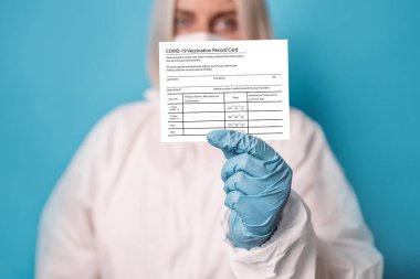 Koruyucu giysi maskeli doktor. Yeni COVID-19 Aşı Kartı 'nı gösteriyor. Parlak mavi arkaplanda Corona virüsü aşısı var.