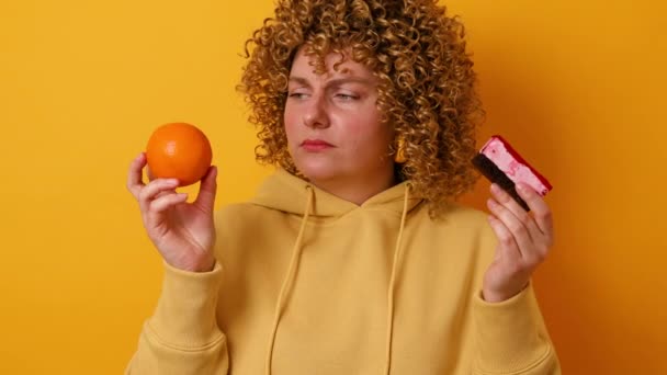 Portré frusztrált tétovázó 30-as nő visel alkalmi jumper kezében édes torta és friss egészséges narancs sárga színű háttér stúdió. Fogyás, sport, fitnesz, diéta koncepció. 4k — Stock videók