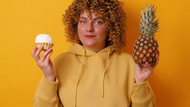 Νεαρή γυναίκα κάνει επιλογή μεταξύ ανανά και νόστιμο γλυκό κέικ απομονώνονται σε κίτρινο χρώμα φόντο. Καλοκαίρι, διατροφή και υγιεινός τρόπος ζωής — Αρχείο Βίντεο