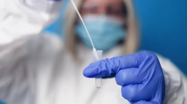 Coronavirus PCR testi. Koruyucu giysi maskeli doktor mavi arka planda teşhis amaçlı tükürük örneği tutan eldiven.