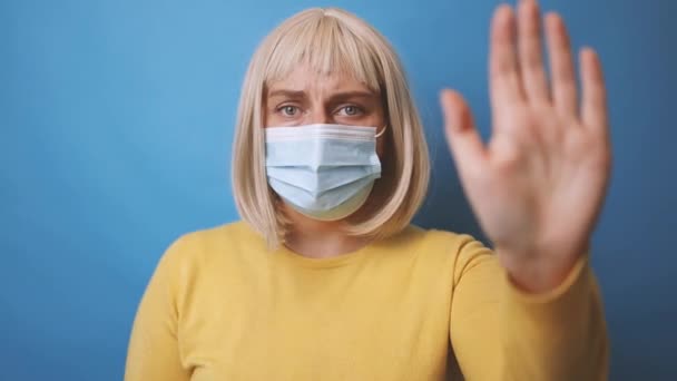 Портрет 30-х років молодої жінки в захисній медичній масці з жовтим светром робить стоп жест рукою над ізольованим синім тлом — стокове відео