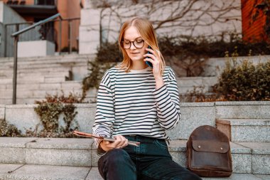 Serbest bir iş. 30 'lu yaşlardaki başarılı sarışın kadın bahar günü gökdelenin yanında otururken açık havada akıllı telefon ve tablet kullanıyor.